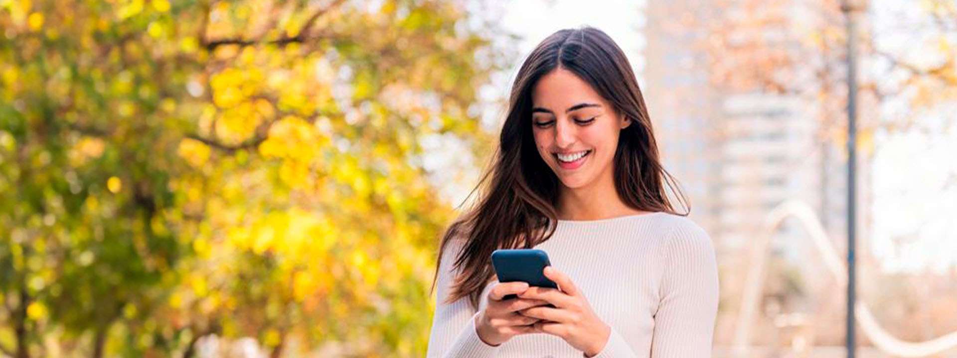 A woman stands outdoors on a sunny day, smiling as she looks at her smartphone, with trees and a blurred cityscape in the background.