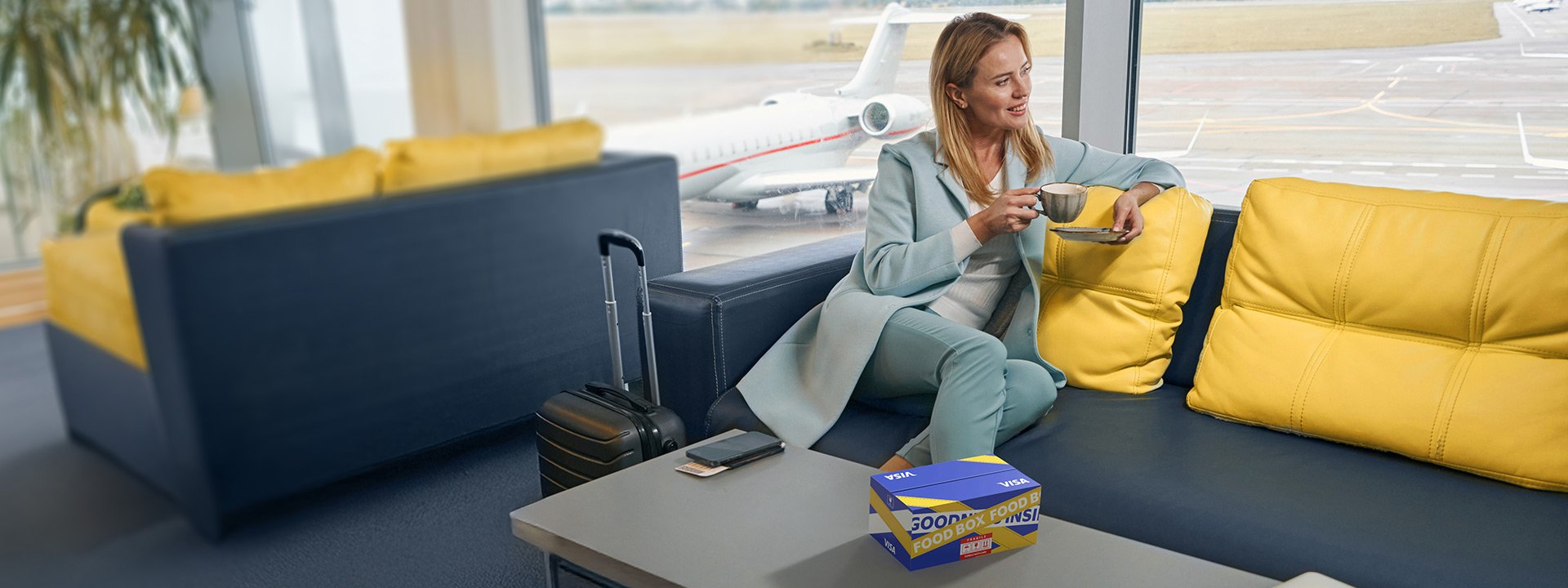 A woman in a light blue outfit sits on a yellow and blue couch at an airport lounge, holding a drink with a snack box nearby. A small suitcase and a tablet are on the table. An airplane is in the background.