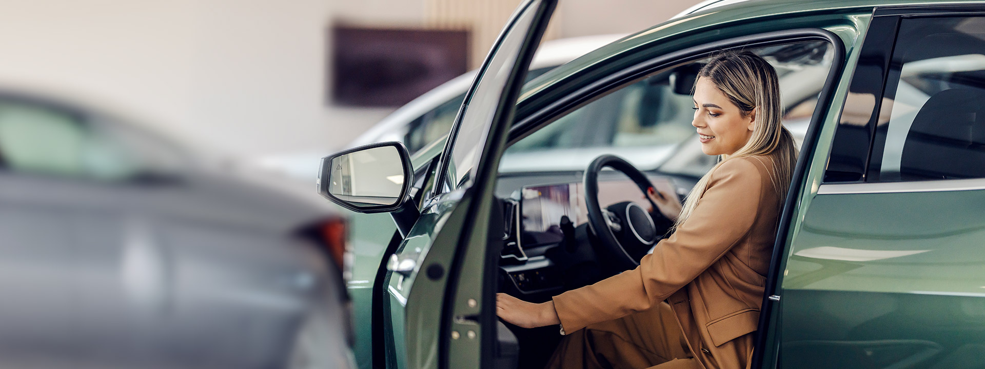 A woman in a tan suit sits in the driver’s seat of a green car with the door open, appearing to inspect the vehicle.