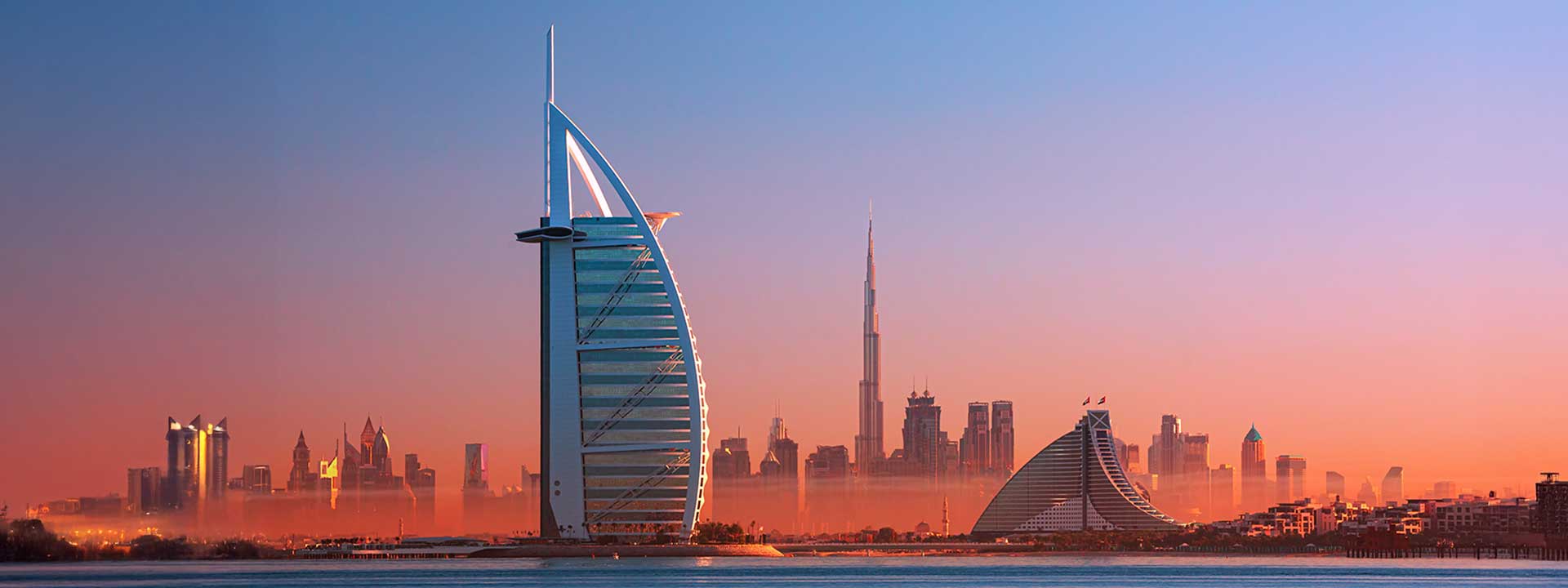 Skyline of Dubai at dusk featuring the Burj Al Arab in the foreground and the Burj Khalifa in the hazy distance, against a gradient sky.