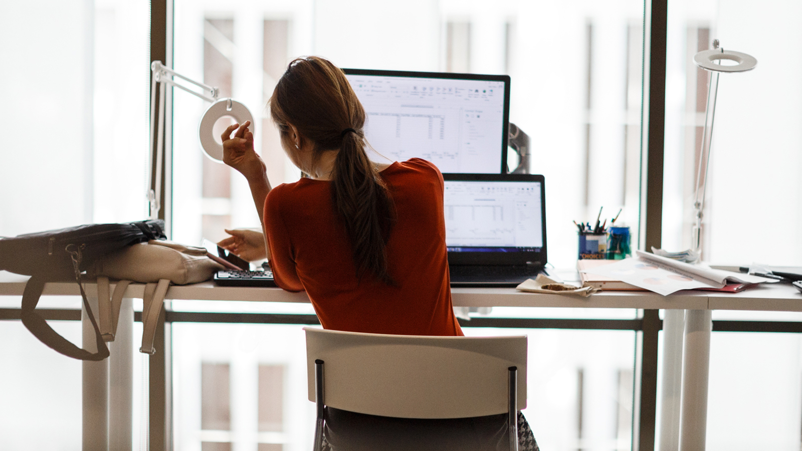 Woman sitting down with a laptop, monitor, books and bags on a desk.