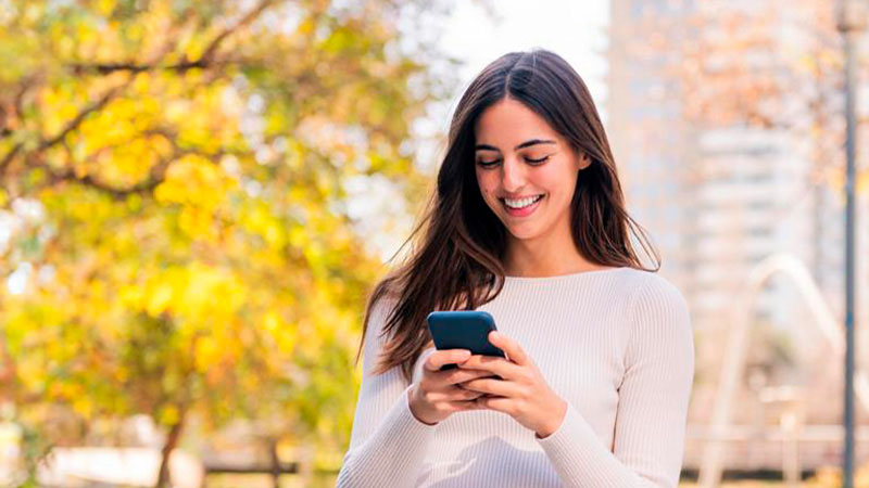 A woman stands outdoors on a sunny day, smiling as she looks at her smartphone, with trees and a blurred cityscape in the background.