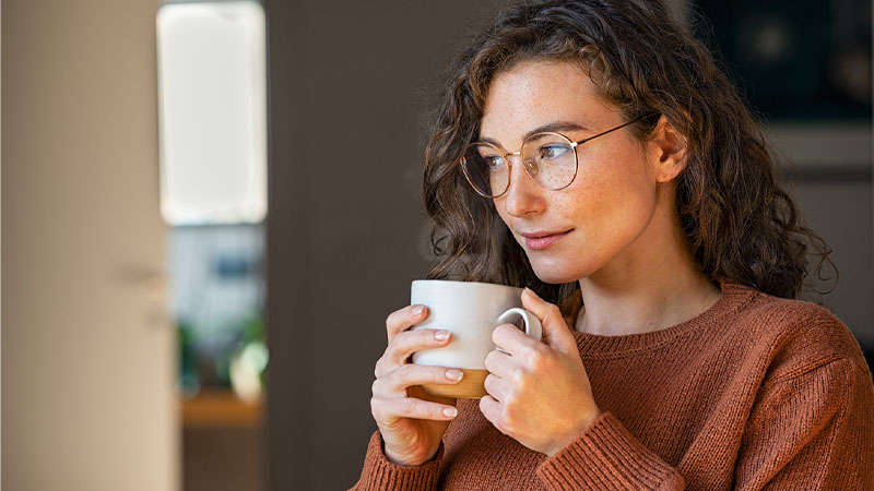 A person in a brown sweater holding a white mug close to their chest, standing indoors with a bright window and room interior in the background.