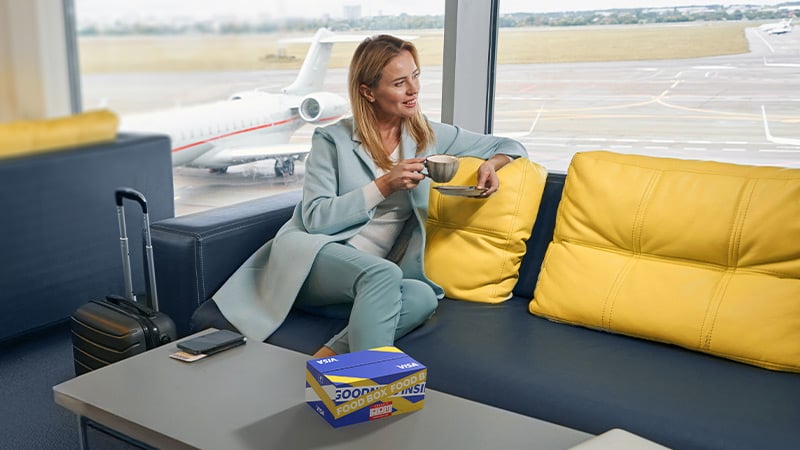 A woman in a light blue outfit sits on a yellow and blue couch at an airport lounge, holding a drink with a snack box nearby. A small suitcase and a tablet are on the table. An airplane is in the background.