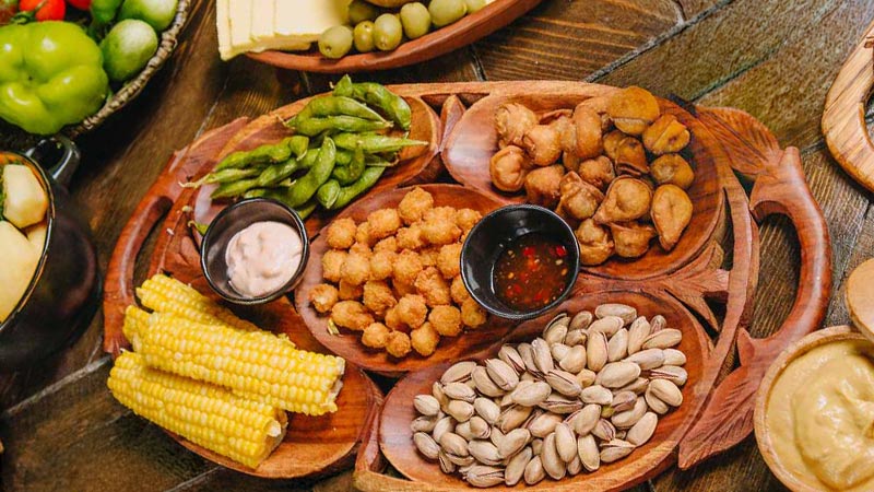 Wooden platter with assorted snacks, corn, nuts, and dipping sauces.