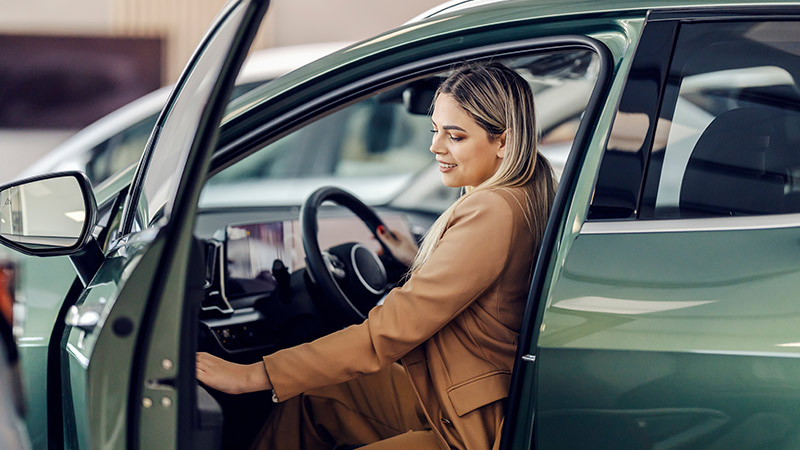 A woman in a tan suit sits in the driver’s seat of a green car with the door open, appearing to inspect the vehicle.