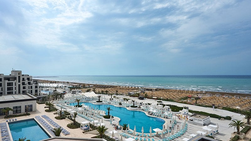 Aerial view of a beachfront resort featuring outdoor swimming pools, lounge chairs, palm trees, and rows of umbrellas on the sandy shore beside the sea.