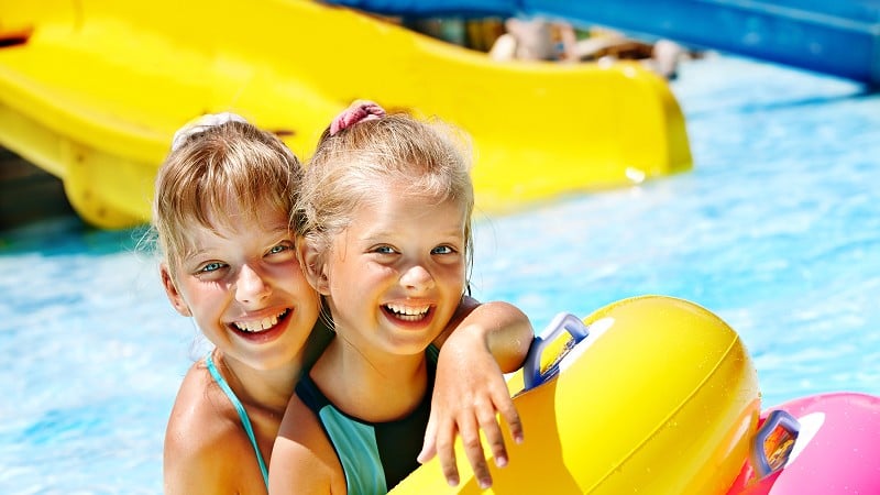 Two young girls smiling in a pool, holding onto a yellow inflatable raft, with water slides visible in the background.