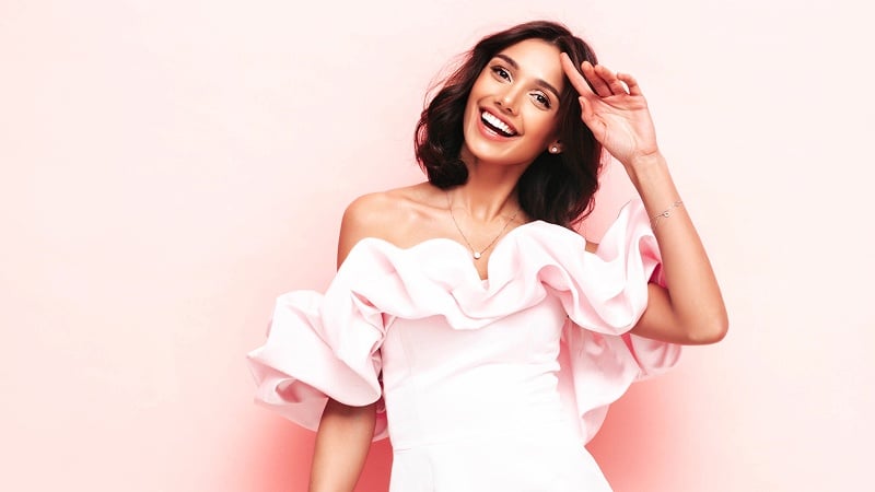 Woman in a white dress with ruffled pink off-the-shoulder sleeves stands smiling against a light pink background.