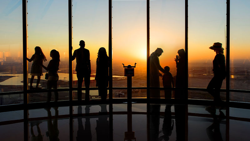 Silhouettes of people stand by large windows, observing a sunset view from a high vantage point inside a building.