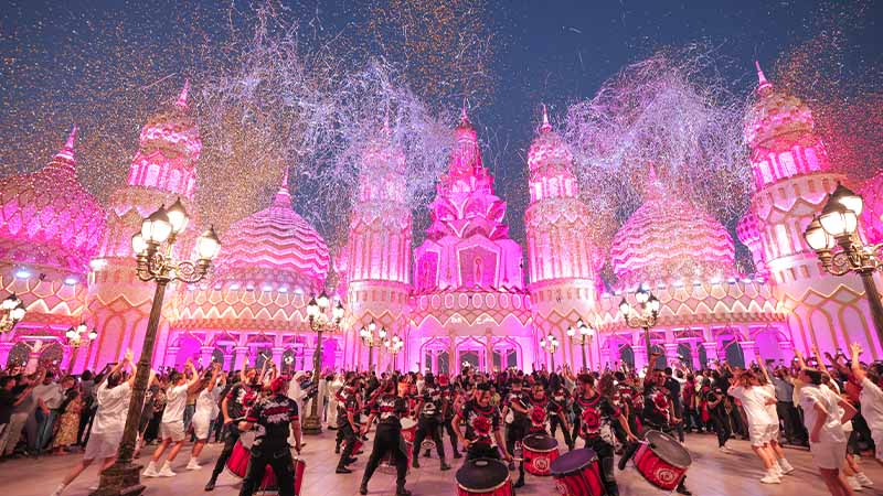A large group of performers dances in front of a brightly lit, ornate pink building with fireworks and confetti in the night sky.