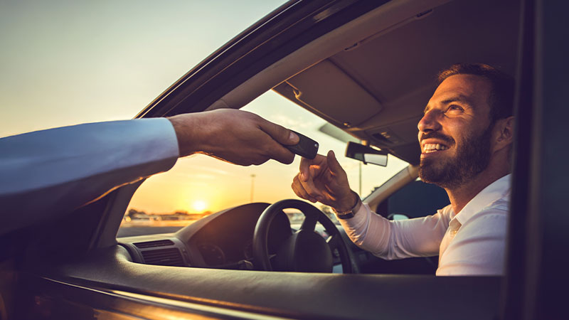 A man in a car receives a key fob from someone outside the vehicle at sunset, smiling through the open window.