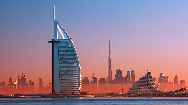 Skyline of Dubai at dusk featuring the Burj Al Arab in the foreground and the Burj Khalifa in the hazy distance, against a gradient sky.