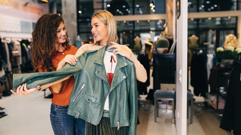 Two women in a store holding a green leather jacket, smiling and discussing the item. Clothing and accessories are displayed in the background.
