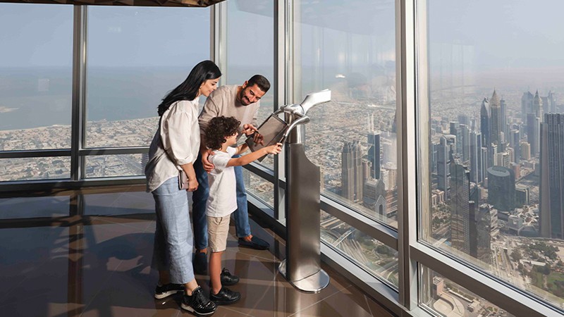 A family looks through a telescope at a cityscape from a high observation deck with large windows.