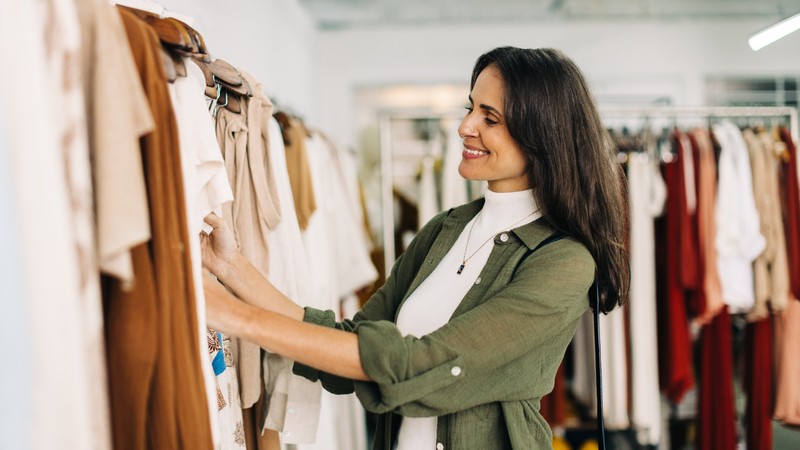 A woman in a green shirt smiles while browsing through clothes on a rack in a store.