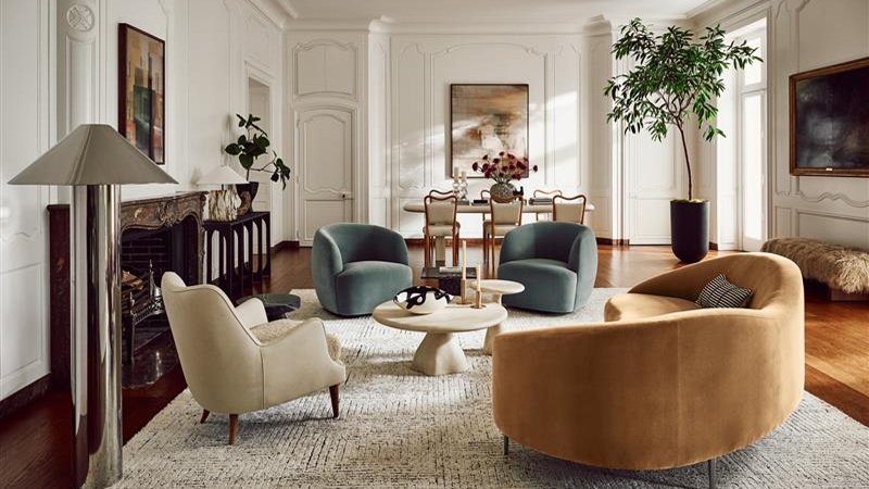 Luxurious living room with modern furniture, including a tan sofa, gray chairs, a round coffee table, and a dining area. Large plant and wall art enhance the stylish ambiance.