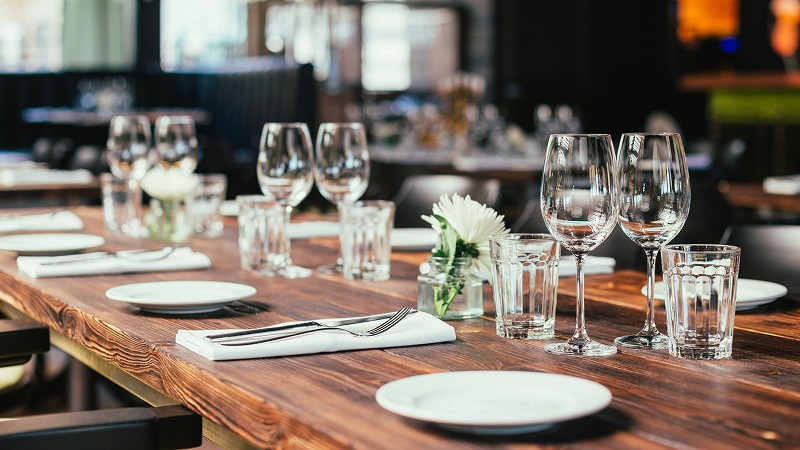 A wooden table set for dining with empty plates, wine glasses, water glasses, and silverware. Small white flowers in vases are placed along the table.