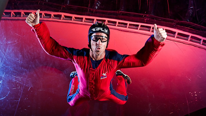 Person in a red flight suit and helmet is indoor skydiving, floating in a vertical wind tunnel with thumbs up, against a red and purple background.