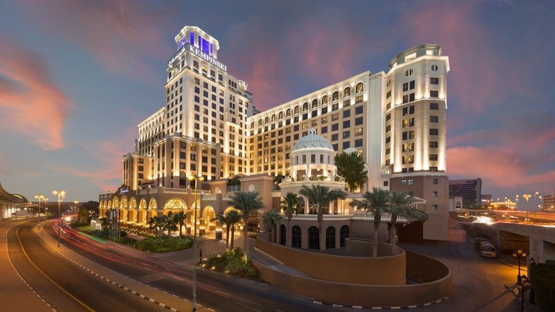 A large, illuminated hotel building at dusk with a dome feature and surrounding palm trees. A road curves alongside, with blurred car lights visible.