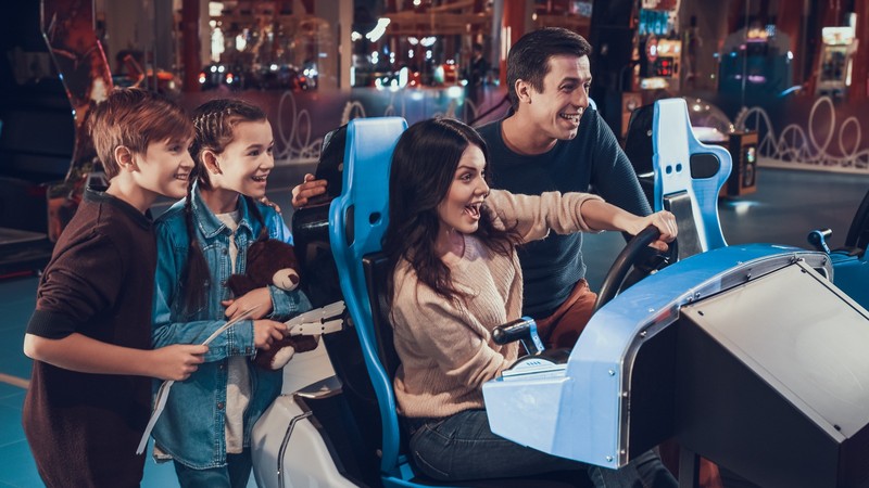 A woman is driving a blue racing arcade game with a man and two children watching and smiling in an amusement arcade.