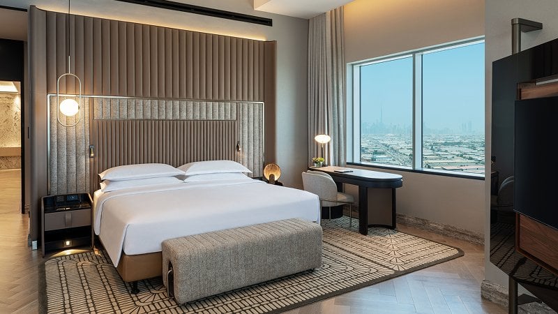 Modern hotel room with a large bed, textured headboard, and a patterned rug. A desk and chair are near a window with a city view. Neutral tones dominate the decor.