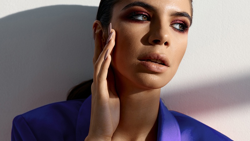 Woman wearing a purple blazer poses against a white wall, touching her face with her hand and looking to the side. Dramatic makeup and shadow are visible.
