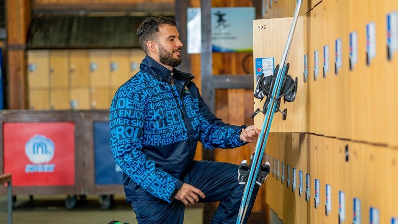 Man in blue ski jacket kneeling to access lockers, with skis beside him.