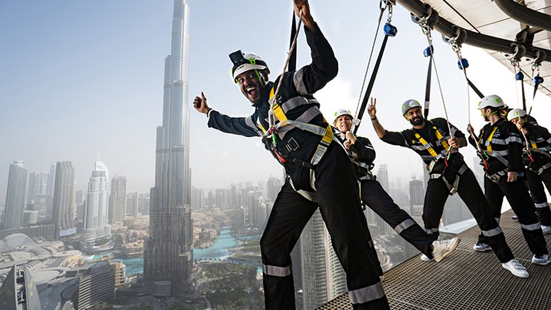 Several people in safety harnesses stand on the edge of a tall building, smiling and posing, with the Burj Khalifa and Dubai city skyline in the background.