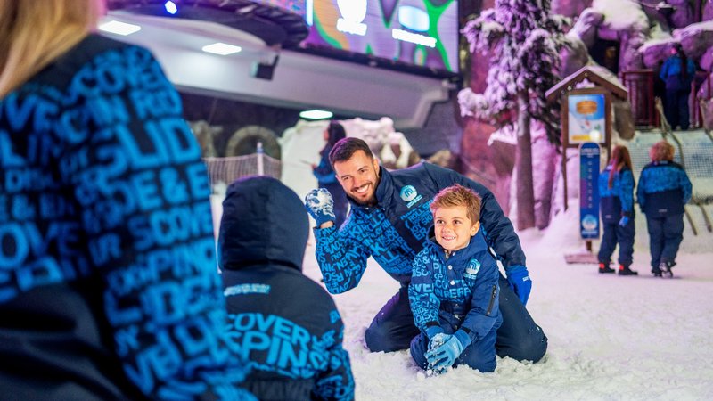 Adults and children wearing snow gear play in an indoor snow park, surrounded by artificial snow and themed decorations.
