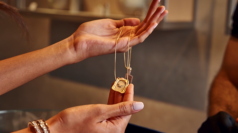 A person holds up a gold chain necklace with a square and circular pendant in a jewelry store.