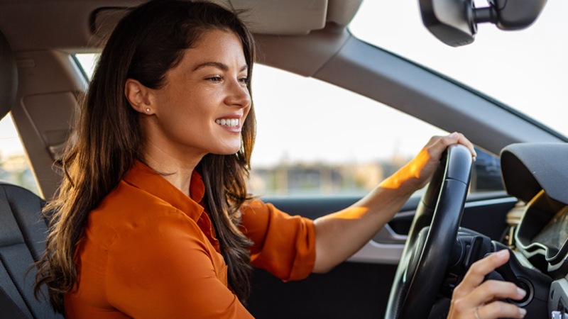A woman in an orange top is smiling while driving a car, with sunlight illuminating the interior.