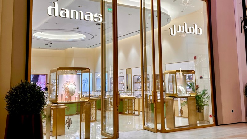 A jewelry store with large glass windows displays jewelry inside glass cases. The store sign reads "Damas" in both Latin and Arabic scripts.