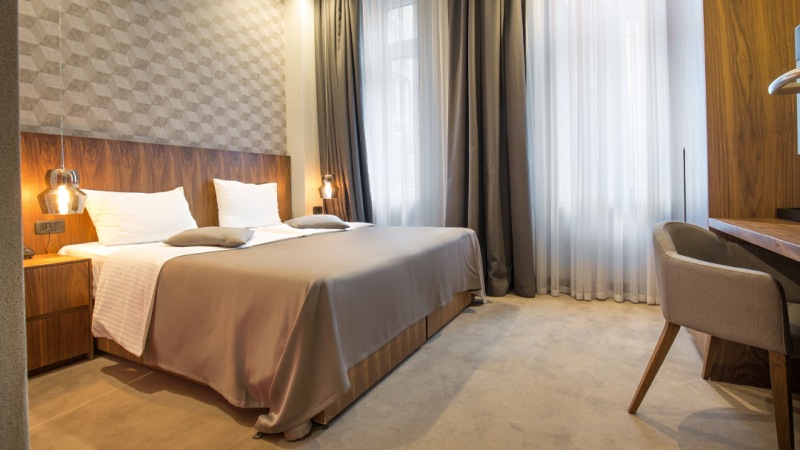 A modern hotel room with a neatly made bed, two bedside lamps, and a desk with a chair. Light enters through large windows with sheer and heavy curtains.