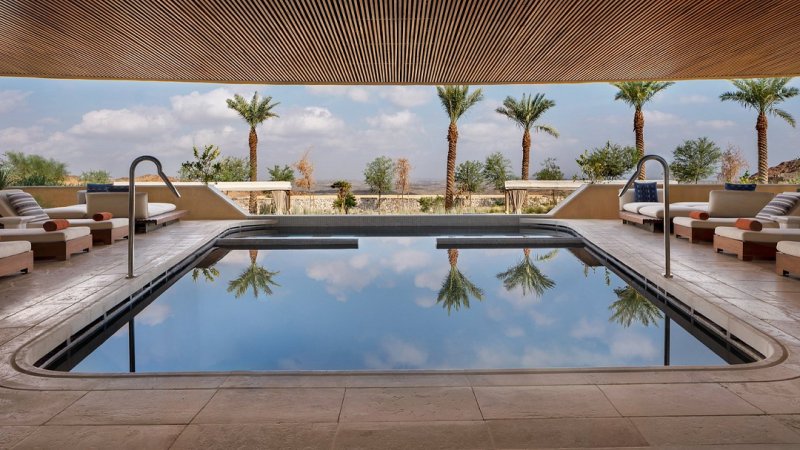 An indoor pool with a skylight reflects the cloudy sky, surrounded by lounge chairs. Palm trees are visible outside.