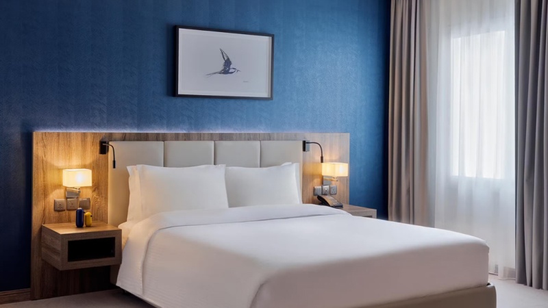 Hotel room with a white bed, blue accent wall, bird artwork, two bedside tables with lamps, and a window with sheer curtains.