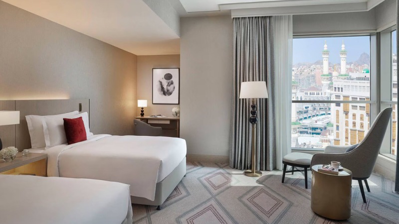 Hotel room with two beds, a desk, and a chair near a window offering a city view. Neutral color palette with modern decor and geometric patterned carpet.