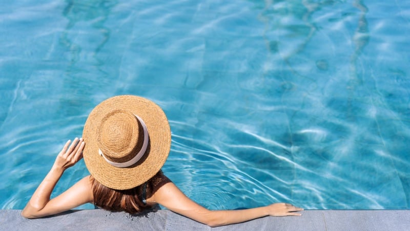 Person with a straw hat lounging at the edge of a swimming pool with clear blue water.