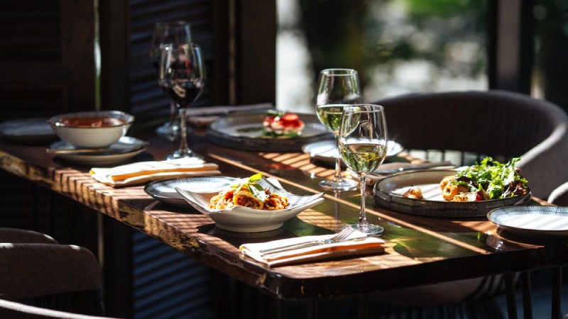 A sunlit table set for four with plates of food, wine glasses, and napkins.