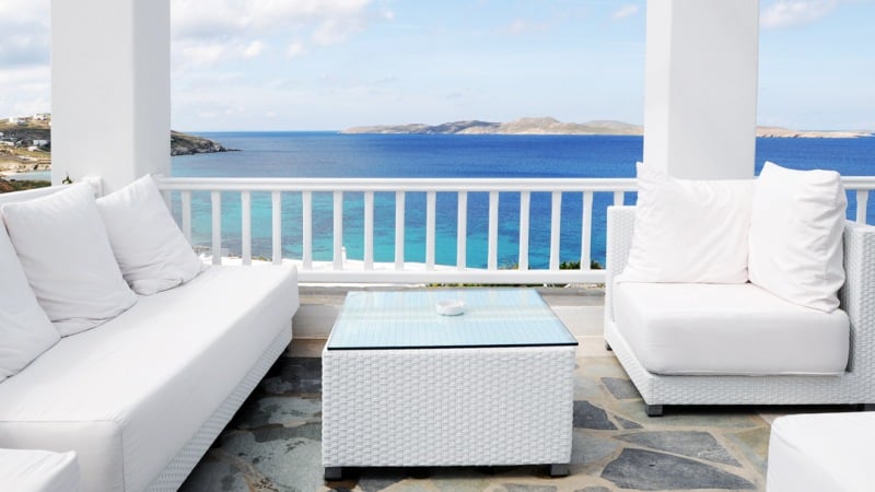 White patio furniture on a stone-floored terrace with a glass-top table, overlooking a blue sea and a distant coastline under a cloudy sky.
