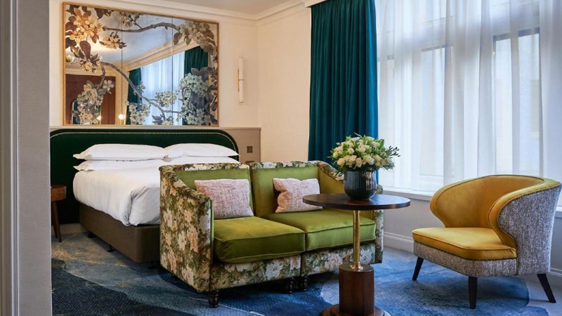 Hotel room with a neatly made bed, floral-patterned couch, round table with a flower vase, and an upholstered chair. A large mirror with floral details hangs above the bed.